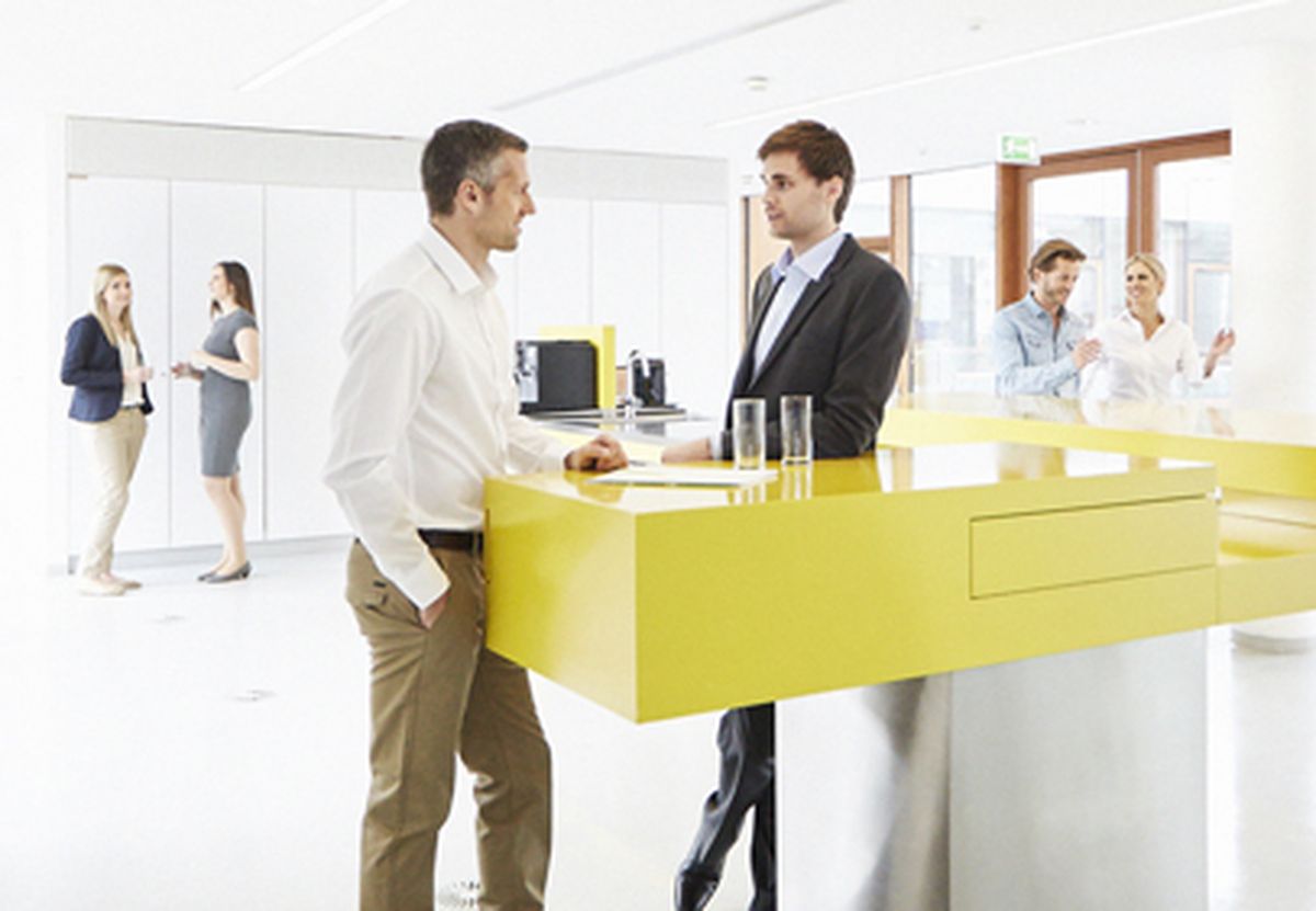 Six KEBA employees are standing in pairs around a yellow coffee counter, deeply engaged in conversation.