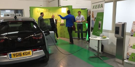Stand espositivo di KEBA e Loxone con una BMW i3 in carica presso una wallbox. Persone discutono davanti allo sfondo verde della sezione "Smart Home".