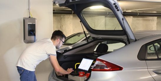 Un uomo analizza dei dati con un laptop dal bagagliaio aperto di un'auto elettrica collegata a una stazione di ricarica KEBA in un garage sotterraneo.